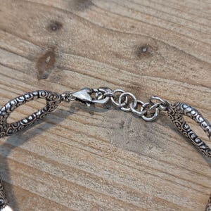 K&ouml;nnte beinhalten: Nahaufnahme eines silberfarbenen Halskettenverschlusses und einer Kette. Die Kette hat ein verziertes, gemustertes Design. Der Verschluss hat ein herzf&ouml;rmiges Design. Die Halskette liegt auf einer Holzoberfl&auml;che.