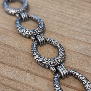 K&ouml;nnte beinhalten: Nahaufnahme einer silberfarbenen Kette mit ovalen Gliedern. Jedes Glied ist mit einem aufwendigen, wirbelnden Muster verziert. Die Kette wird auf einer Holzoberfl&auml;che pr&auml;sentiert, wodurch das detaillierte Design und der metallische Glanz des Schmucks hervorgehoben werden.