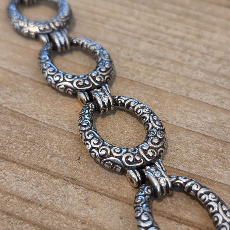 K&ouml;nnte beinhalten: Nahaufnahme einer silberfarbenen Kette mit ovalen Gliedern. Jedes Glied ist mit einem aufwendigen, wirbelnden Muster verziert. Die Kette wird auf einer Holzoberfl&auml;che pr&auml;sentiert, wodurch das detaillierte Design und der metallische Glanz des Schmucks hervorgehoben werden.