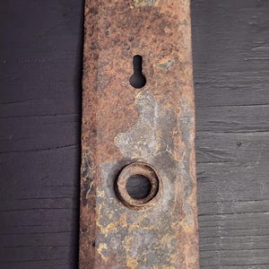 May include: A rectangular metal door plate featuring a heavy layer of brown and orange rust. The plate includes a central circular opening for a knob, a keyhole opening above it, and four small circular screw holes located at the corners.