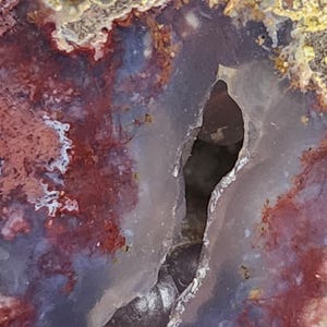 May include: Close-up of a polished stone with a cavity. The stone features a mix of colours, including deep reds, purples, and hints of yellow and grey. The cavity is dark and elongated, adding depth to the natural stone.