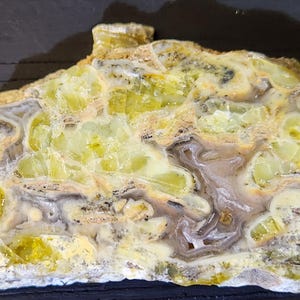 May include: A cross-section of a mineral specimen, showcasing a blend of colors and textures. The stone features a mix of pale yellow, cream, and gray hues, with intricate patterns and crystalline structures. The overall appearance suggests a natural, raw gemstone.