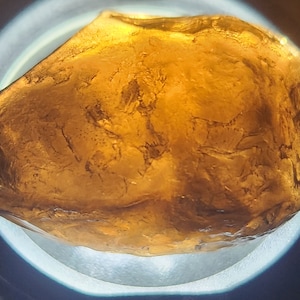 Op de afbeelding: Een grote, onregelmatig gevormde, amberkleurige edelsteen met een glad, gepolijst oppervlak. De steen is doorschijnend en vertoont interne strepen.