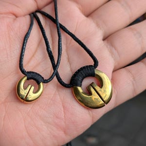 May include: Two gold necklaces with black cords and geometric pendant charms. The larger pendant is a circle with a split in the middle. The smaller pendant is a smaller circle with a split in the middle.