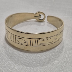 Pode incluir: Uma pulseira de metal em tom dourado com acabamento polido e um padrão geométrico gravado composto por diamantes e linhas verticais. A pulseira tem um design aberto com um mecanismo de fecho circular de gancho na parte superior.