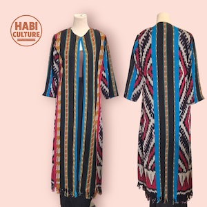 Peut inclure: Longue veste ouverte à rayures colorées noires, bleues, rouges et jaunes. Le vêtement a des manches trois-quarts et un ourlet à franges. Le logo "HABI CULTURE" est visible dans le coin supérieur gauche.