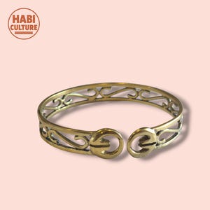 Puede incluir: Brazalete de metal dorado con un diseño abierto. El brazalete presenta intrincados adornos y un cierre de broche. El logotipo de la marca "HABI CULTURE" está en la esquina superior izquierda.