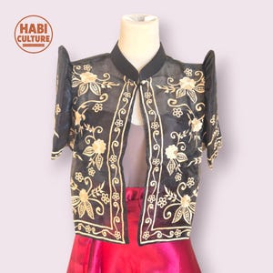 May include: Black bolero jacket with floral embroidery in gold thread. The jacket has short sleeves and a mandarin collar. The garment is displayed on a mannequin, paired with a red skirt. The logo "HABI CULTURE" is visible in the upper left corner.