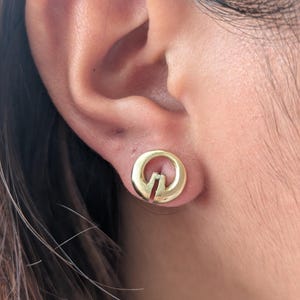May include: A gold-coloured stud earring with a circular design and a vertical slit. The earring is worn on an ear, with the skin tone appearing light. The hair is brown with highlights.