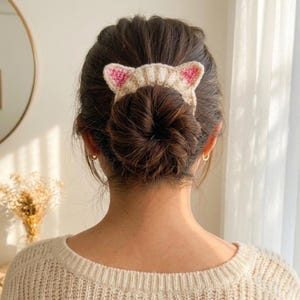 Puede incluir: Un accesorio para el cabello de ganchillo beige con forma de orejas de gato, con detalles rosas en el interior. El accesorio para el cabello se lleva en un mo&ntilde;o. La persona lleva un su&eacute;ter color crema.