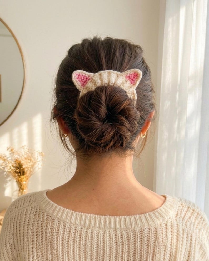 Puede incluir: Un accesorio para el cabello de ganchillo beige con forma de orejas de gato, con detalles rosas en el interior. El accesorio para el cabello se lleva en un mo&ntilde;o. La persona lleva un su&eacute;ter color crema.