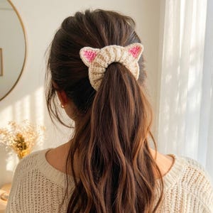 Puede incluir: Una goma para el pelo con forma de orejas de gato, de color beige y rosa, se lleva en una coleta. La goma para el pelo est&aacute; hecha de material de punto. Las orejas de gato son rosas y la goma para el pelo es beige. La persona lleva un jersey color crema.