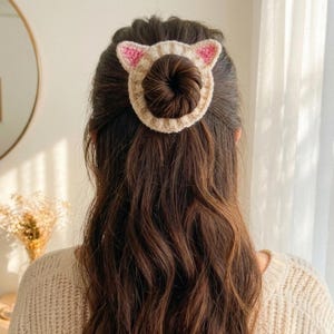 Puede incluir: Un recogedor de pelo de ganchillo beige con forma de cara de gato, con detalles rosas en las orejas. El accesorio para el pelo se lleva en el pelo, con el pelo casta&ntilde;o oscuro del usuario visible. Un bonito accesorio para el pelo.