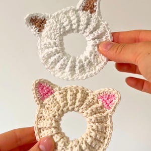 Puede incluir: Dos coleteros hechos a mano con forma de gato de ganchillo. Uno es blanco con detalles de orejas marrones, el otro es beige con detalles de orejas rosas. Los coleteros son circulares y tienen una textura acanalada.