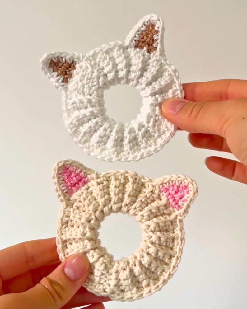 Puede incluir: Dos coleteros hechos a mano con forma de gato de ganchillo. Uno es blanco con detalles de orejas marrones, el otro es beige con detalles de orejas rosas. Los coleteros son circulares y tienen una textura acanalada.