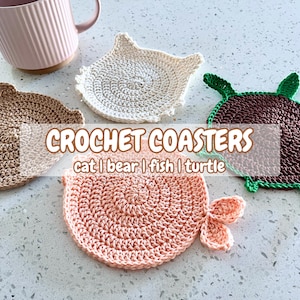 May include: Crocheted coasters in cat, bear, fish, and turtle shapes. The coasters are in shades of brown, white, and pink, with green accents on the turtle. A pink mug is in the background. The text "CROCHET COASTERS" is visible.