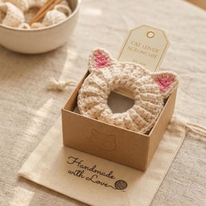 Puede incluir: Una scrunchie beige con forma de gato, tejida a ganchillo, con detalles de orejas rosas, presentada en una peque&ntilde;a caja marr&oacute;n. La caja est&aacute; sobre una bolsa con cord&oacute;n de color crema con las palabras "Handmade with Love" impresas. Una etiqueta dice "Cat Lover Scrunchie".