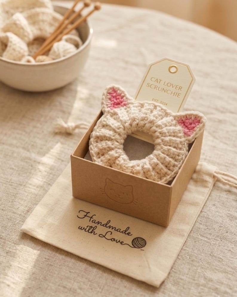 Puede incluir: Una scrunchie beige con forma de gato, tejida a ganchillo, con detalles de orejas rosas, presentada en una peque&ntilde;a caja marr&oacute;n. La caja est&aacute; sobre una bolsa con cord&oacute;n de color crema con las palabras "Handmade with Love" impresas. Una etiqueta dice "Cat Lover Scrunchie".