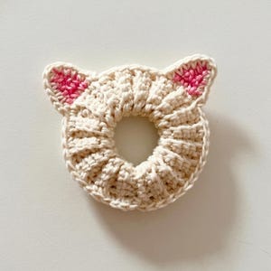 Puede incluir: Una goma para el pelo de ganchillo de color crema con orejas de gato. Las orejas est&aacute;n acentuadas con detalles rosas. La goma para el pelo es circular con un agujero en el centro, y el punto de ganchillo crea una apariencia texturizada.