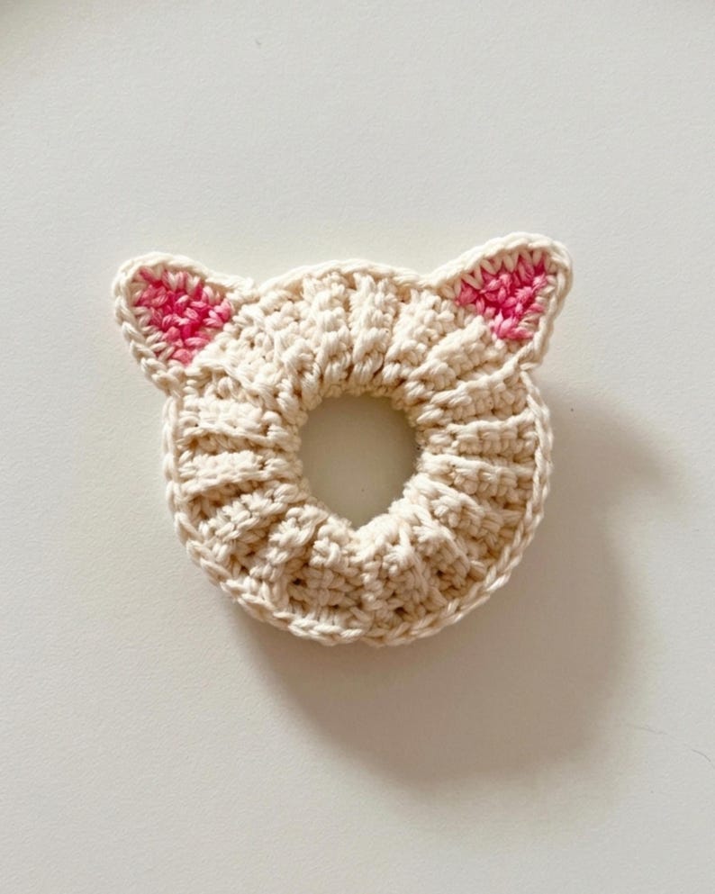 Puede incluir: Una goma para el pelo de ganchillo de color crema con orejas de gato. Las orejas est&aacute;n acentuadas con detalles rosas. La goma para el pelo es circular con un agujero en el centro, y el punto de ganchillo crea una apariencia texturizada.