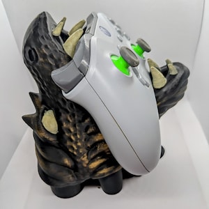 May include: A gray and white Xbox controller rests in a black and gold monster-themed controller holder. The holder has sharp teeth and a textured surface. The controller has green accents on the joysticks.