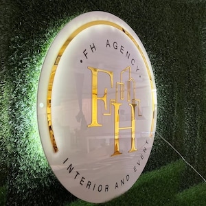 Può includere: Un'insegna rotonda e illuminata con la scritta "FH AGENCY. INTERIOR AND EVENT" in oro. L'insegna ha uno sfondo bianco con un bordo dorato ed è posta su uno sfondo verde strutturato.