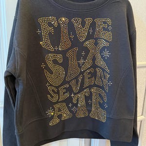 May include: Black sweatshirt with a cropped cut and a large gold rhinestone design that reads "FIVE SIX SEVEN ATE".