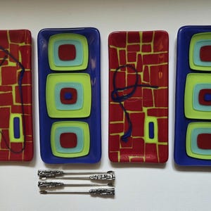 May include: Four rectangular glass serving dishes in red, blue, and green. Two dishes have a red brick pattern, and two have layered squares. Three silver serving forks are below the dishes.