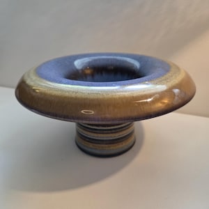 May include: A ceramic pedestal bowl with a wide, flat rim and a central opening. The rim is blue, transitioning to a tan and brown exterior. The base is a cylindrical stack of horizontal stripes in various colors. The bowl is likely used for decorative purposes.