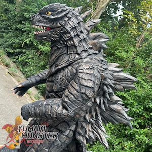 May include: A detailed sculpture of Godzilla, featuring textured, dark gray scales and sharp dorsal fins. The creature has a menacing expression with visible teeth and yellow eyes. The logo "YUMAKI MONSTER STUDIO" is visible.