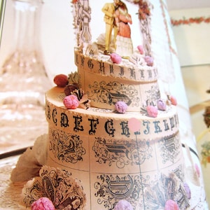 May include: A two-tiered, decorative cake with a vintage design. The cake is adorned with black lettering, floral patterns, and small pink embellishments. A miniature couple stands atop the cake under an archway. The cake is set on a white lace doily.