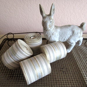 Puede incluir: Cuatro rollos de cinta rayada blanca y dorada en una bandeja de rejilla de alambre con una figurita de conejo de cerámica blanca.