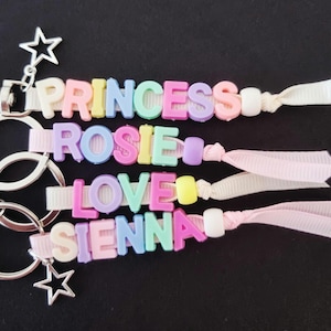 May include: Three personalised keychains with names spelled out in colourful letter beads. The names are "PRINCESS", "ROSIE", "LOVE", and "SIENNA". Each keychain has a silver clasp, star charms, and a ribbon.
