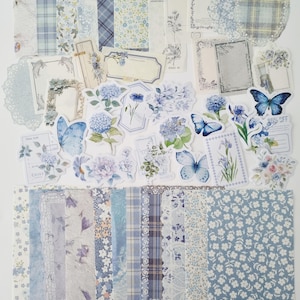 Op de afbeelding: Een verzameling blauw en wit scrapbookpapier, stickers en versieringen. De set bevat papieren met patronen met bloemen, ruiten en kant, samen met stickers met vlinders en bloemen. Het kleurenpalet is zacht en pastel.