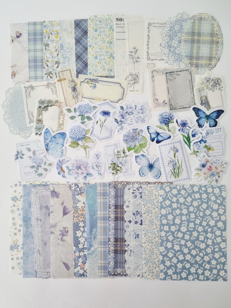 Op de afbeelding: Een verzameling blauw en wit scrapbookpapier, stickers en versieringen. De set bevat papieren met patronen met bloemen, ruiten en kant, samen met stickers met vlinders en bloemen. Het kleurenpalet is zacht en pastel.