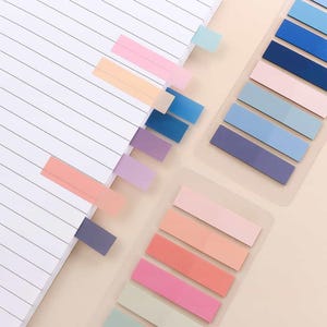 May include: A collection of colorful sticky note tabs in various pastel shades, including pink, peach, blue, and purple. Some tabs are inserted into the pages of a lined notebook, while others are arranged in clear plastic holders. The tabs are rectangular and provide a simple way to mark pages.