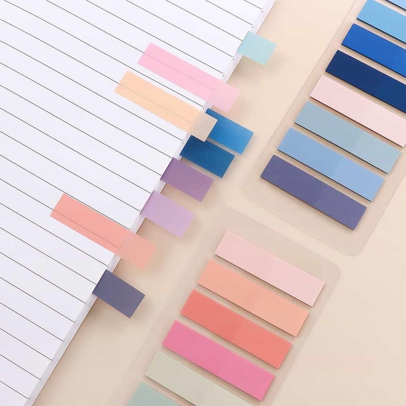 May include: A collection of colorful sticky note tabs in various pastel shades, including pink, peach, blue, and purple. Some tabs are inserted into the pages of a lined notebook, while others are arranged in clear plastic holders. The tabs are rectangular and provide a simple way to mark pages.