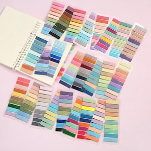 May include: A spiral-bound notebook with lined pages is surrounded by packs of colourful sticky note flags. The flags are arranged in various colour palettes, including pinks, blues, greens, and yellows. The background is a soft pink.