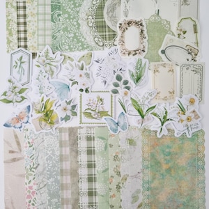 Op de afbeelding: Een verzameling groen en wit scrapbookpapier en uitgestanste versieringen. Het papier heeft bloemen-, ruit- en kanten patronen. Uitgestanste onderdelen zijn onder andere vlinders, bloemen en frames, voor knutsel- en journalingprojecten.