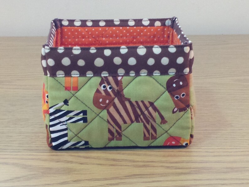 May include: A brown and green fabric storage bin with a polka dot pattern. The bin features a quilted design with animal illustrations, including a zebra, a giraffe, and a horse.