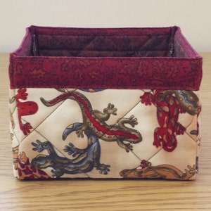 May include: A square, quilted fabric storage bin with a red and white floral pattern. The bin features a red trim and a white background with a repeating pattern of lizards.