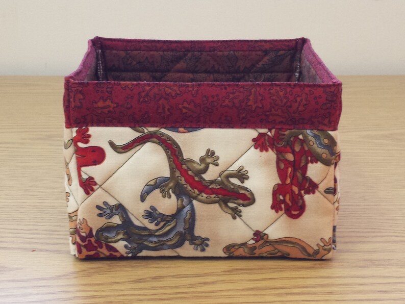 May include: A square, quilted fabric storage bin with a red and white floral pattern. The bin features a red trim and a white background with a repeating pattern of lizards.