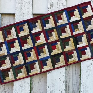 May include: A rectangular table runner with a log cabin quilt pattern in shades of red, blue, green, brown, and cream. The runner is edged with a red border.