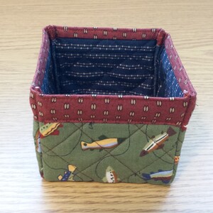 May include: A small, square fabric basket with a quilted design. The exterior features a green fabric with a fish pattern, while the interior has a blue and white dotted fabric. The top edge is trimmed with a maroon fabric.