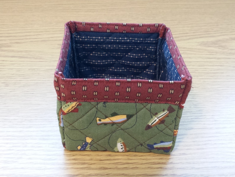 May include: A small, square fabric basket with a quilted design. The exterior features a green fabric with a fish pattern, while the interior has a blue and white dotted fabric. The top edge is trimmed with a maroon fabric.