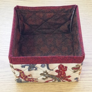 May include: A square fabric storage box with a red trim and a quilted pattern featuring a white background with brown, red, and blue lizards.