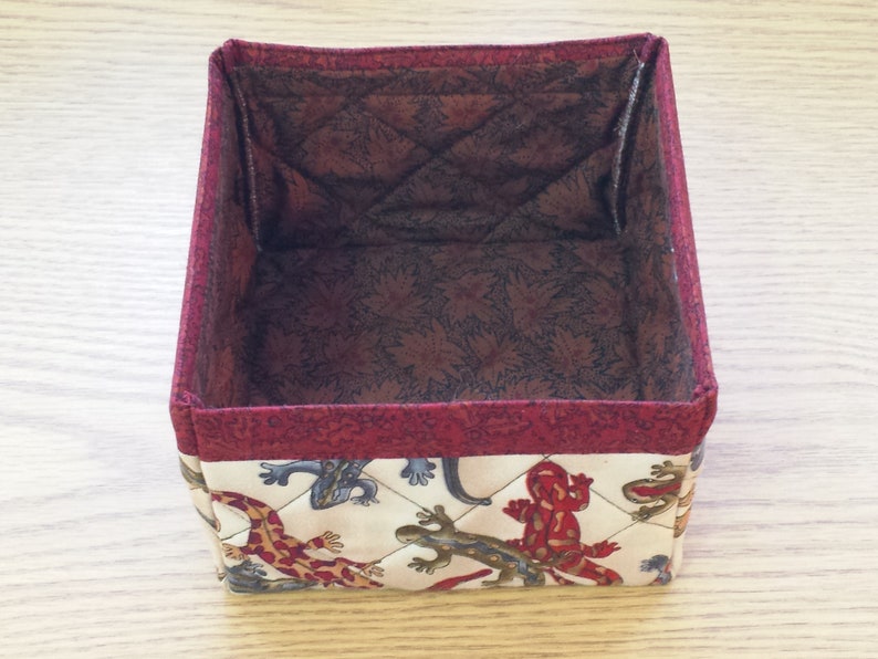 May include: A square fabric storage box with a red trim and a quilted pattern featuring a white background with brown, red, and blue lizards.