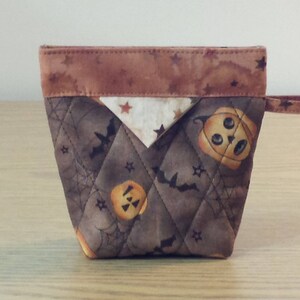 May include: A brown and tan quilted fabric Halloween-themed zippered pouch with a brown fabric handle. The pouch is decorated with a pattern of bats, stars, and pumpkins.