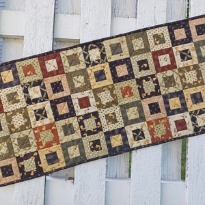 May include: A quilted table runner with a patchwork design featuring squares in shades of brown, green, cream, and red. The squares are arranged in a repeating pattern.