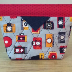 May include: A gray and red quilted fabric pouch with a black flap. The pouch has a repeating pattern of vintage cameras on a gray background.